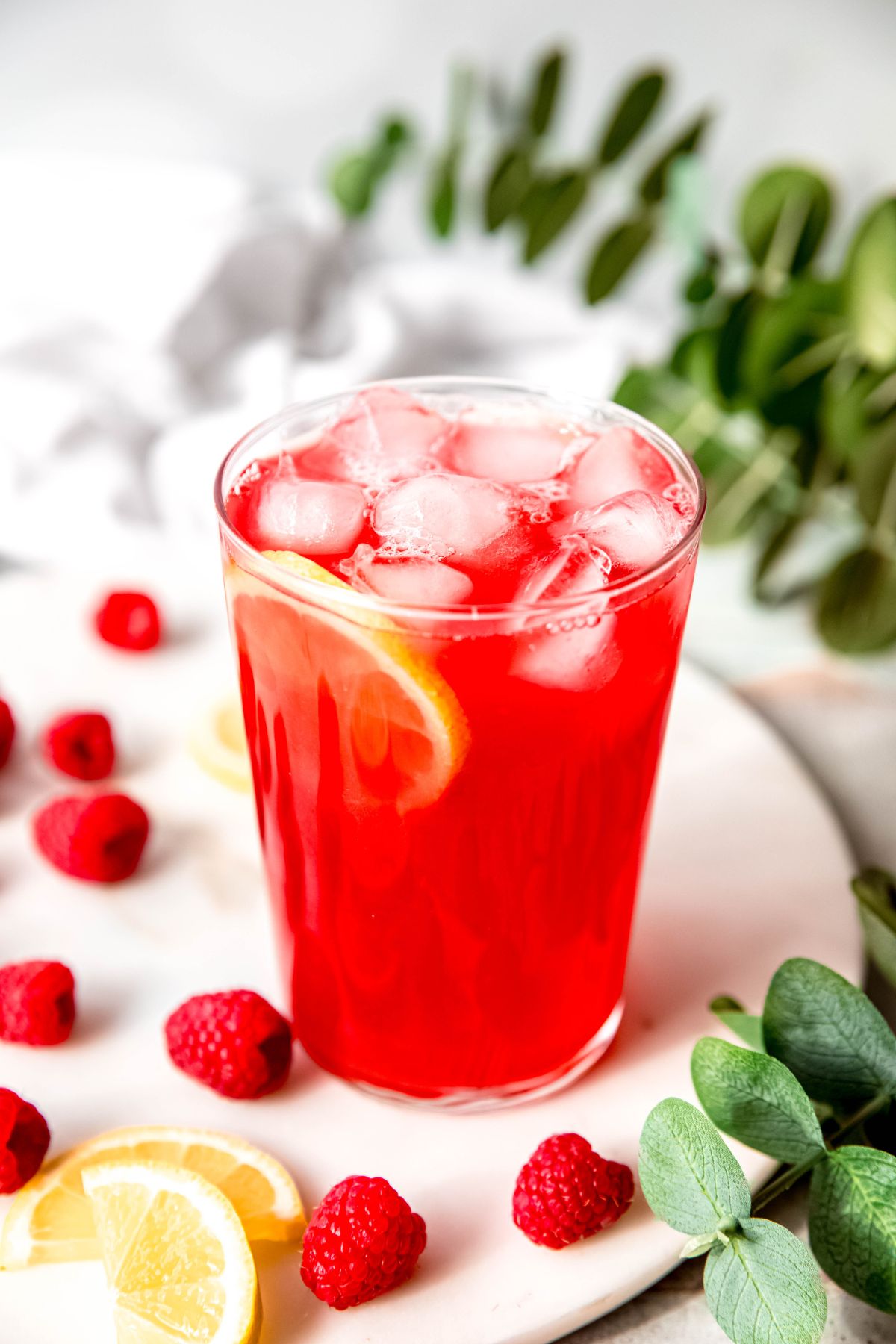 raspberry lemonade in a clear glass on a white table with fresh raspberries and slices of lemon scattered around.