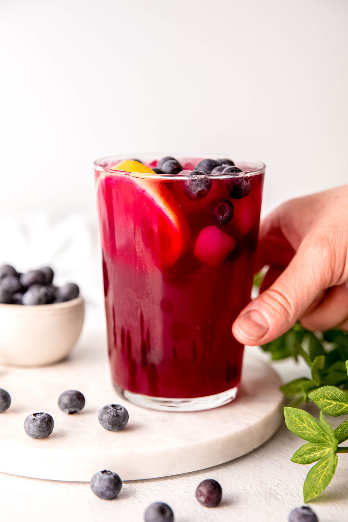 hand grabbing an iced glass of fresh blueberry lemonade from the serving tray with blueberries scattered around on the tray and table.