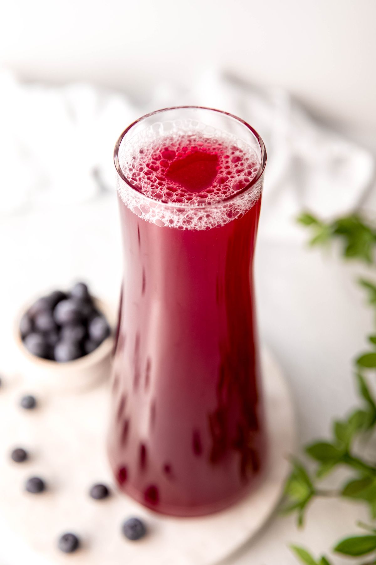 clear handle-less glass pitcher filled with homemade blueberry lemonade on a white table with a small bowl of whole blueberries and fresh berries scattered on the table below.