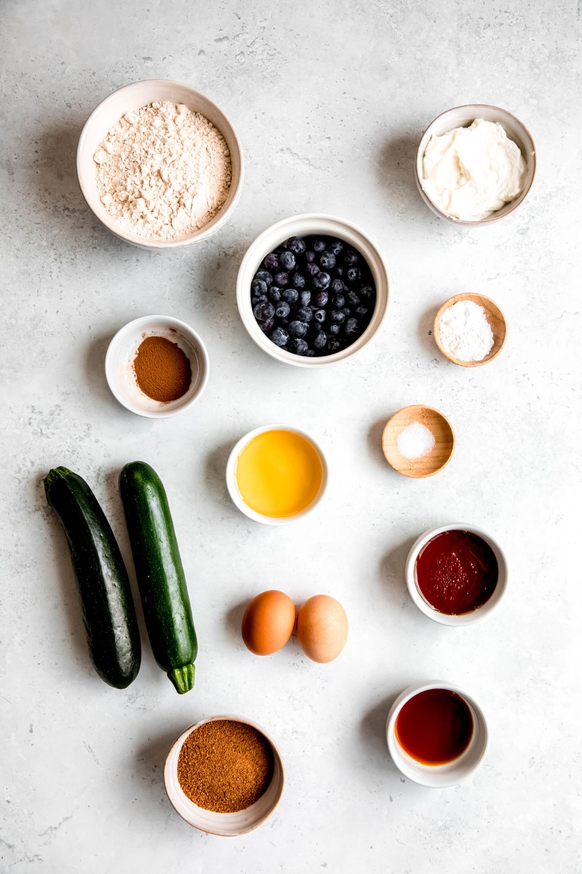 ingredients for making healthy zucchini blueberry muffins measured out into bowls on a white table.