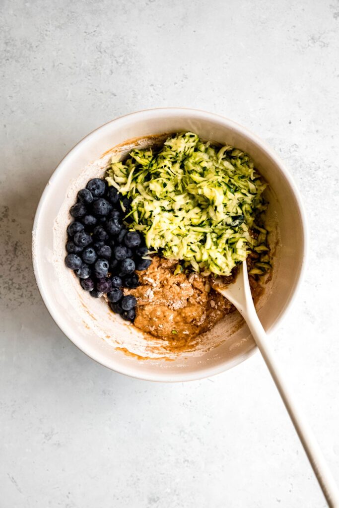 blueberries and shredded zucchini added to the mixing bowl with the dry, chunky looking muffin batter.