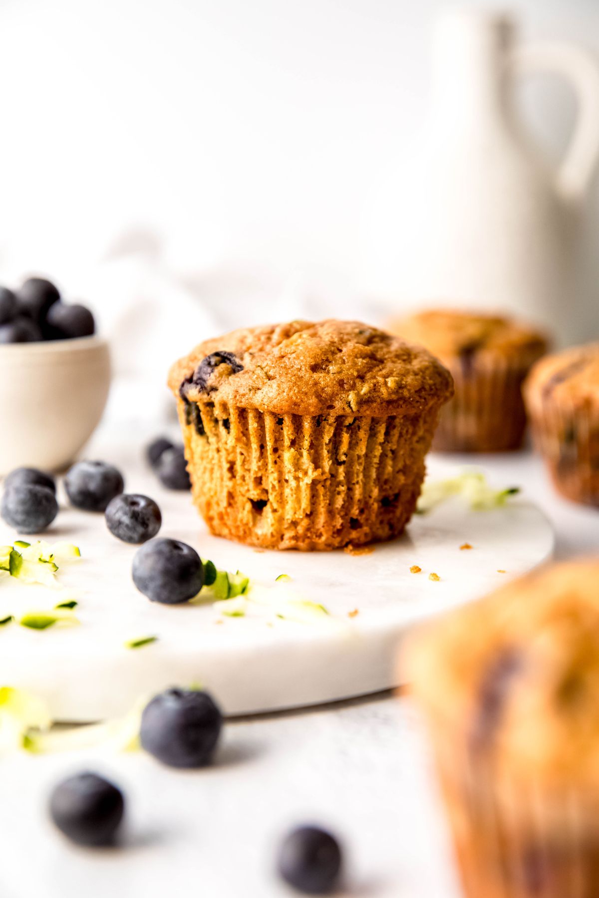 white table with blueberry zucchini muffins, fresh blueberries and zucchini shreds scattered around, and a white bowl of fresh berries.