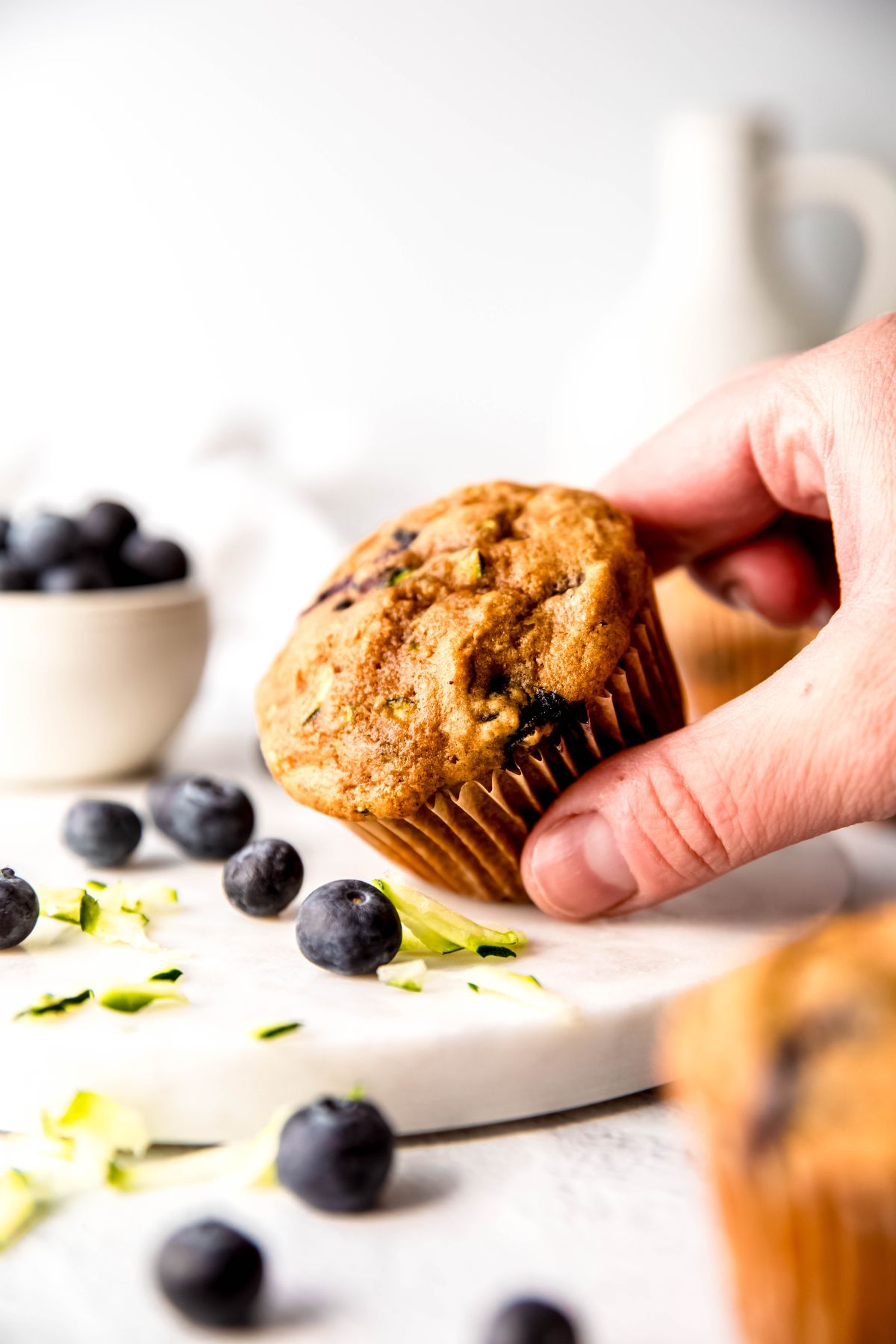 hand grabbing a healthy homemade blueberry zucchini bread muffin from the table, tilting it toward the camera so you can see the golden brown cakey texture with streaks of blueberries and a few visible strands of zucchini.