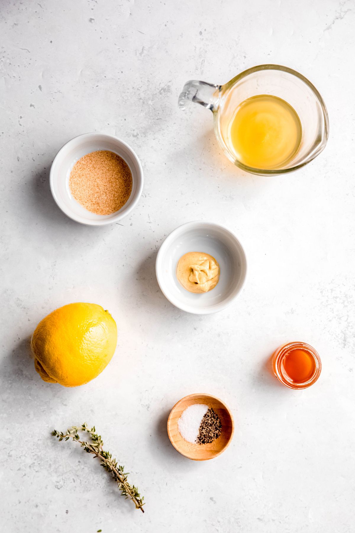 dijon mustard, garlic powder, fresh lemon juice, honey, fresh thyme, salt, and pepper measured out into clear and white glass bowls on a white table to make honey lemon herb vinaigrette.