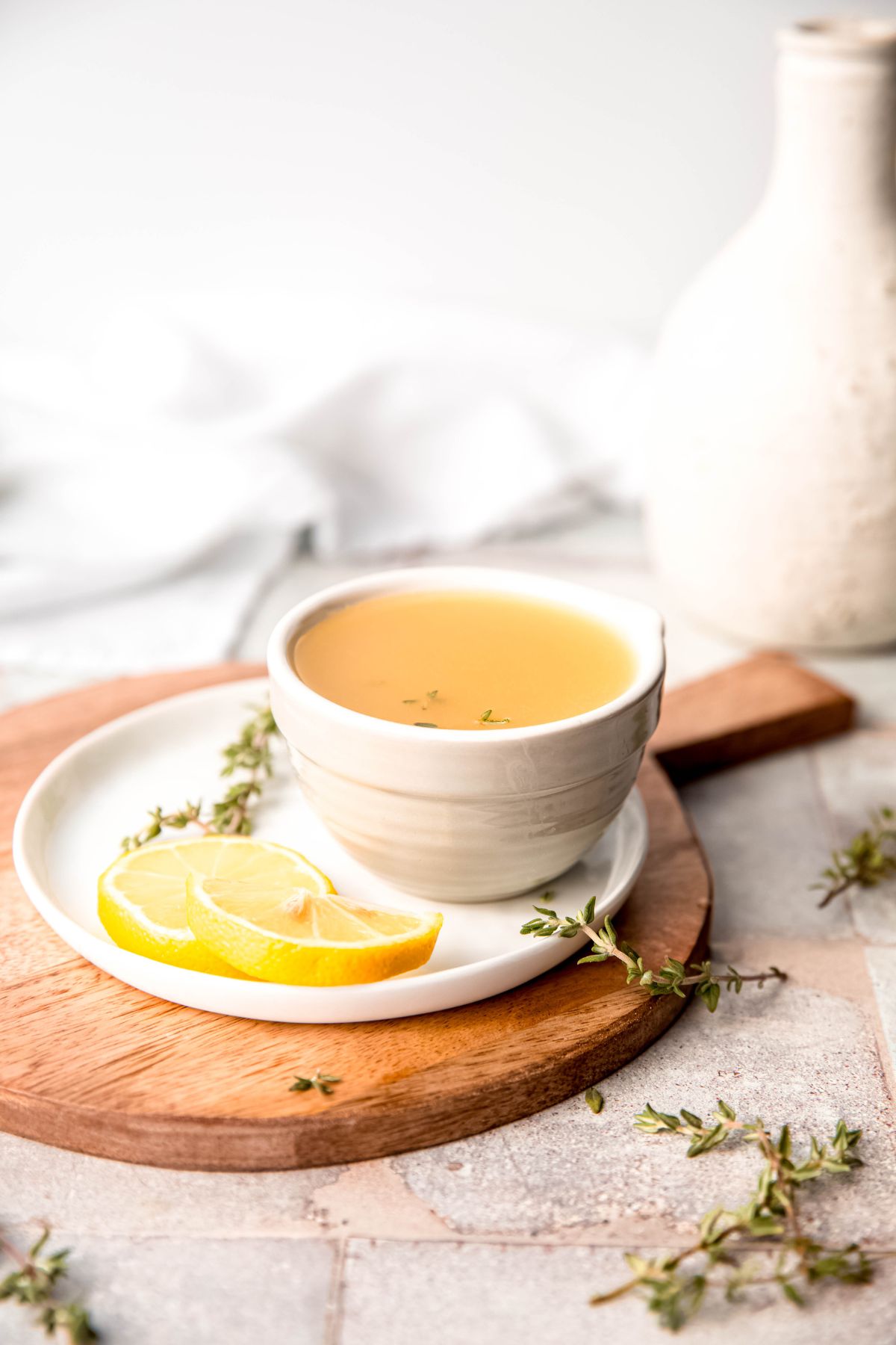 small white bowl of honey dijon lemon vinaigrette with fresh thyme and fresh lemon on a white plate on a wooden serving tray with fresh herb sprigs in the foreground.