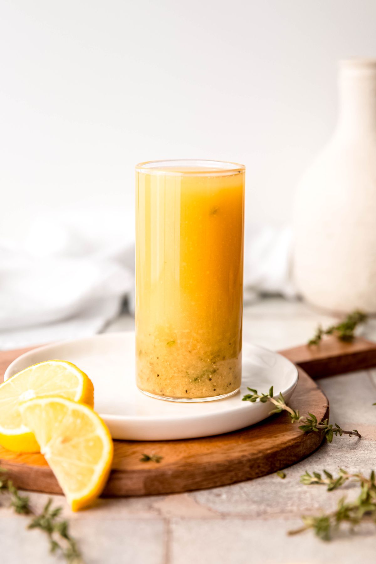 slate grey tile table with a wooden serving tray topped with a white appetizer plate holding a collins glass of homemade lemon honey vinaigrette with a few rounds of fresh lemon and a few sprigs of fresh thyme.