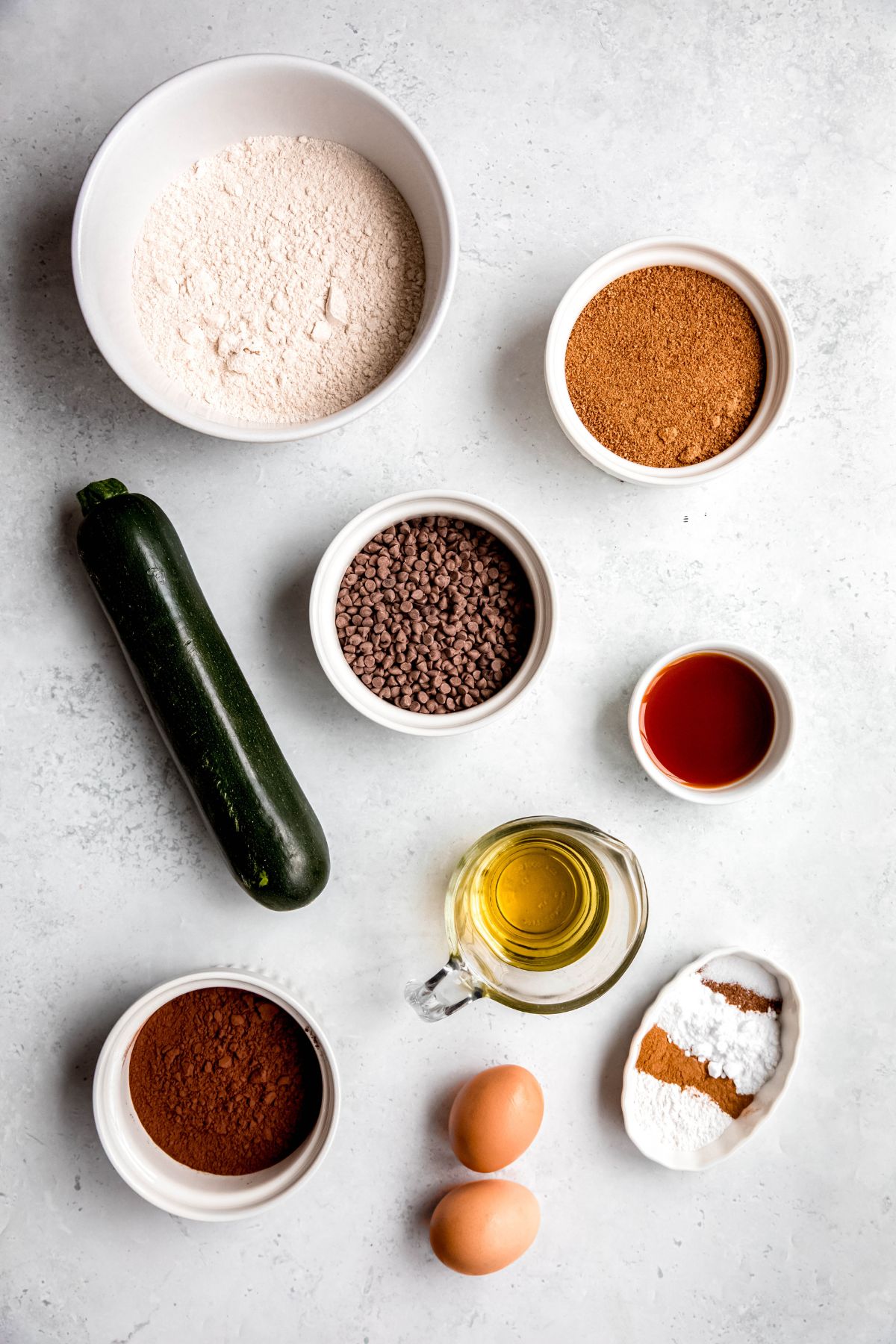 ingredients needed to make healthy chocolate chocolate chip zucchini muffins measured out into bowls on a white table.