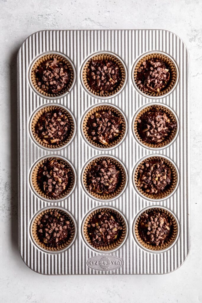 raw chocolate zucchini muffin batter divvied up in the wells of a muffin tin and topped with a sprinkle of mini chocolate chips.