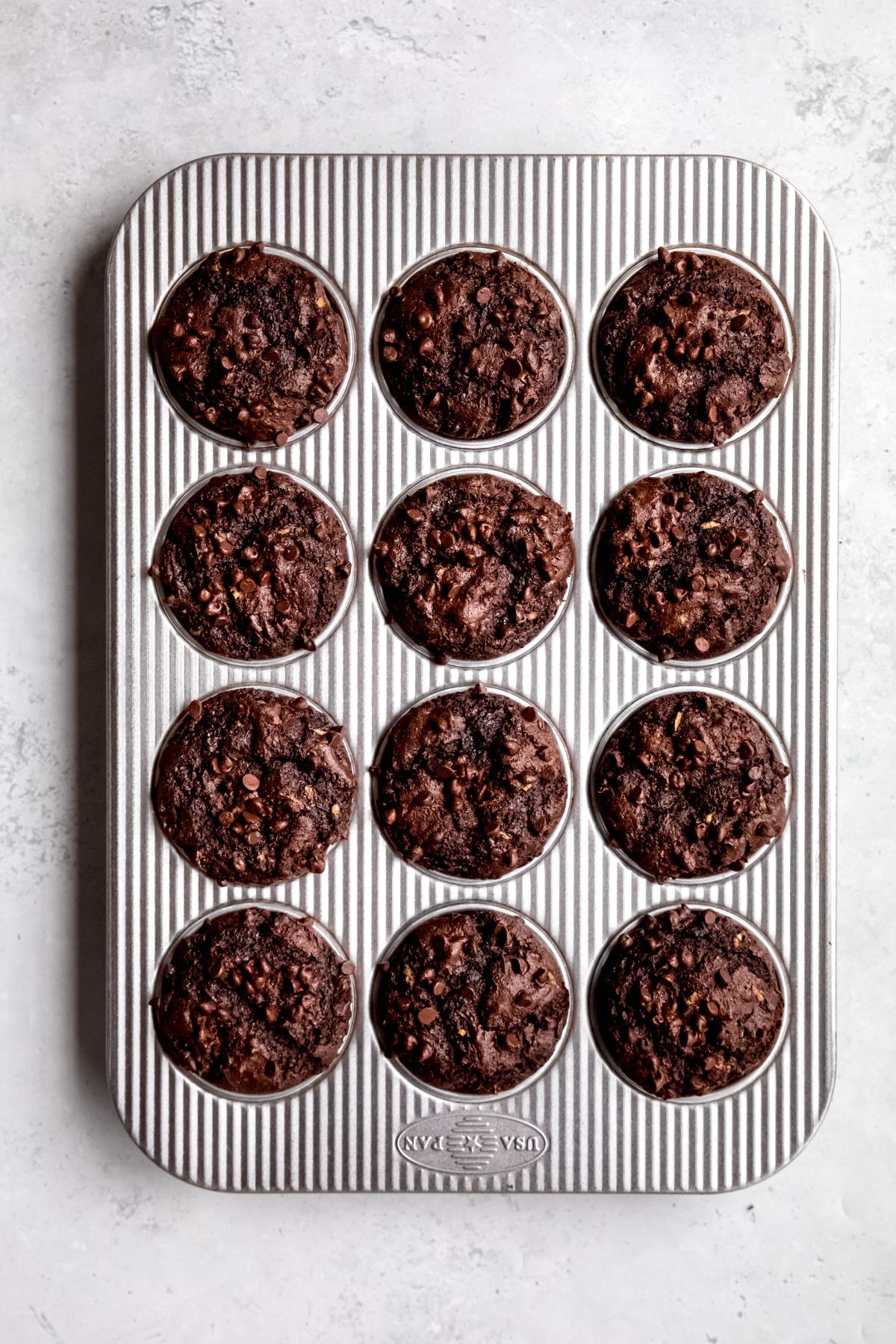 baked double chocolate zucchini muffins in a silver striped muffin tin on a white table.