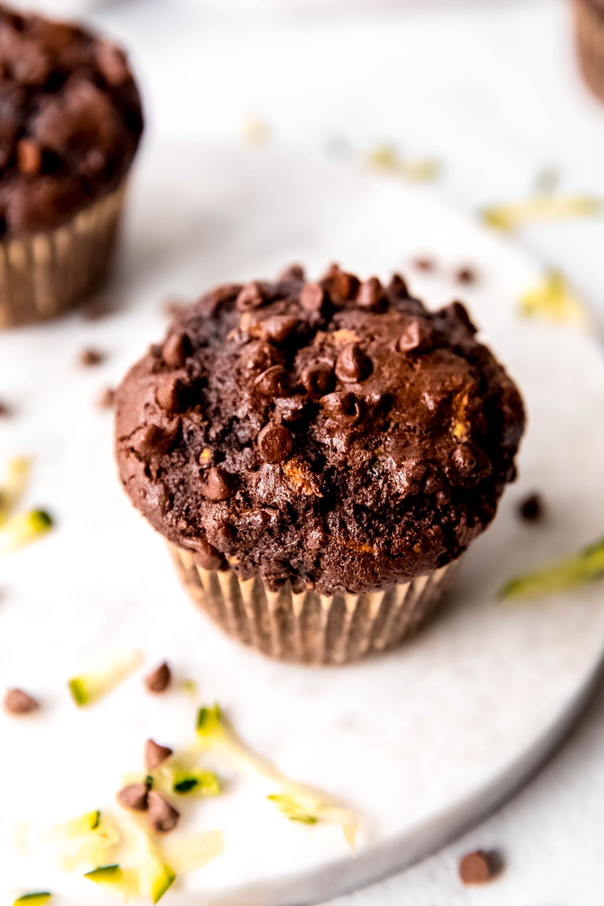closeup 45 overhead shot of the top of a double chocolate zucchini muffin on a white table with mini chocolate chips and shreds of fresh zucchini scattered around.