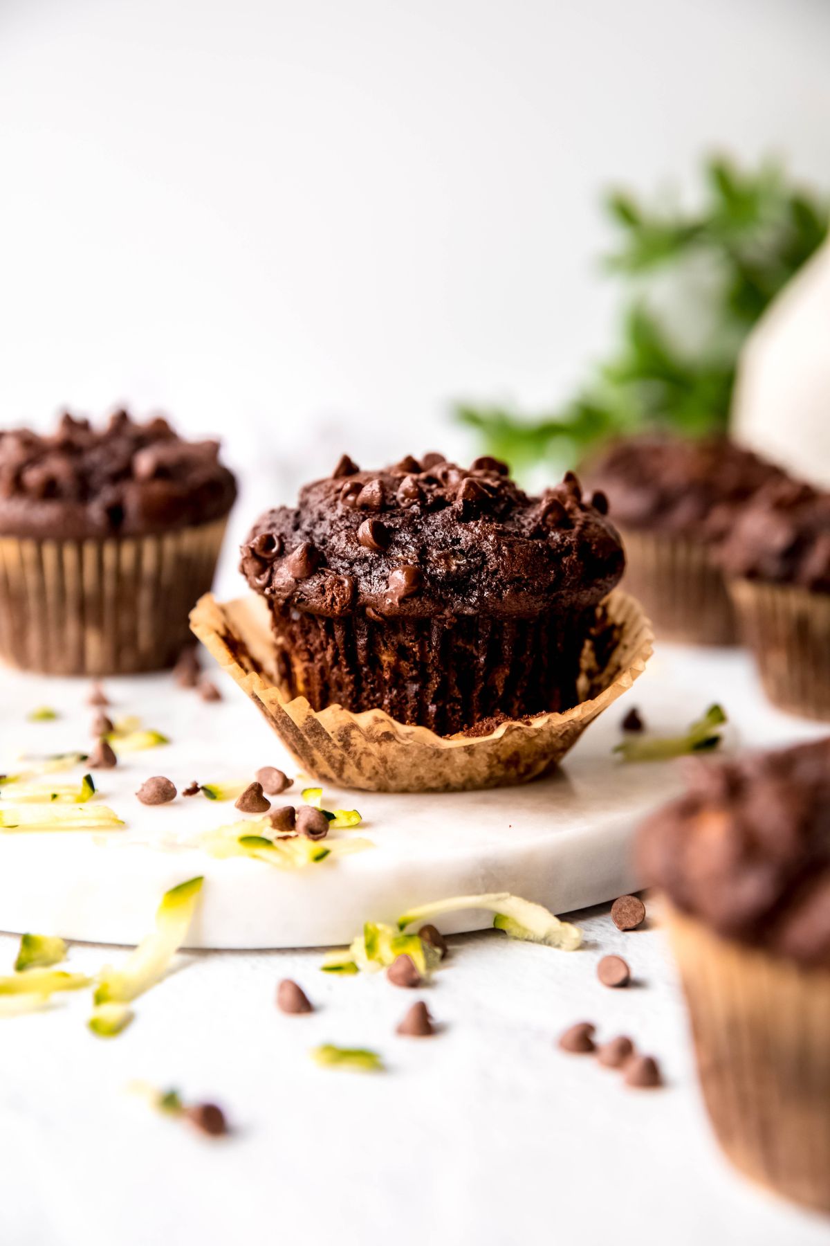 half unwrapped chocolate zucchini chocolate chip muffin on a table with 5 more muffins surrounding it with mini chocolate chips and shredded zucchini sprinkled on the table.