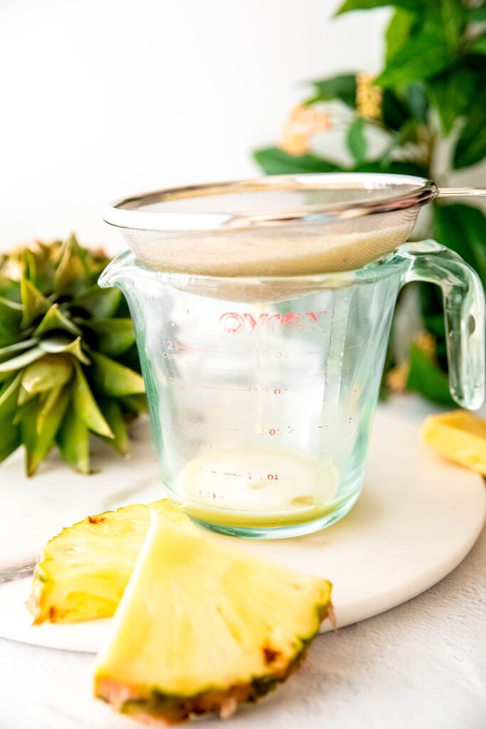 pineapple puree being strained through a mesh strainer into a glass measuring cup.