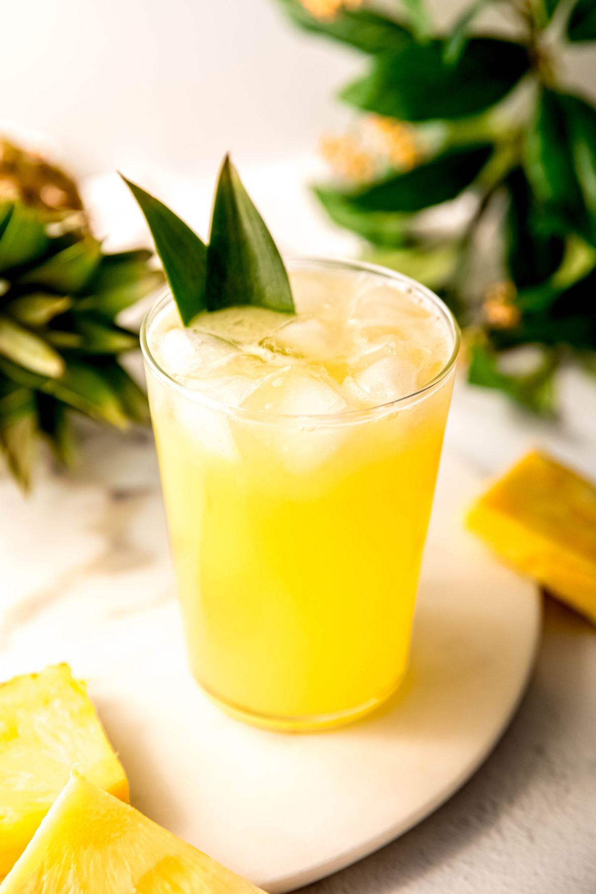glass of pineapple lemonade garnished with pineapple leaves on a white table with fresh pineapple chunks.