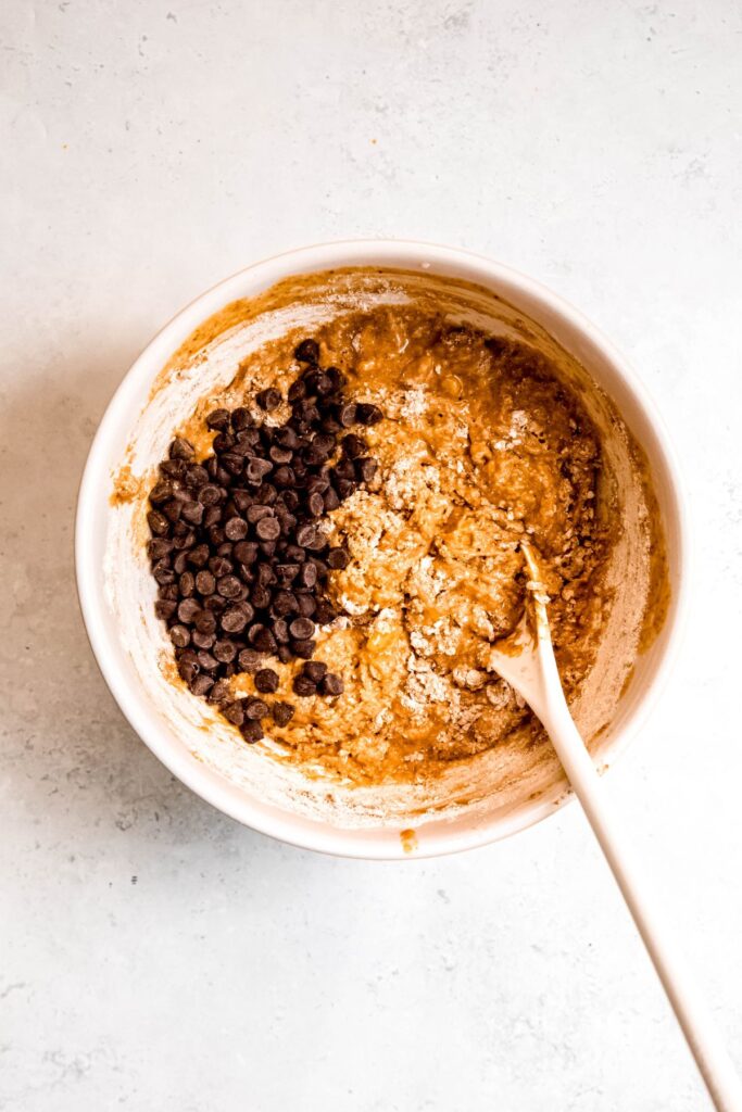protein banana bread batter after mostly stirring in the dry ingredients (there are still visible pockets of flour) in a mixing bowl with a wooden spoon and the chocolate chips added to the bowl.