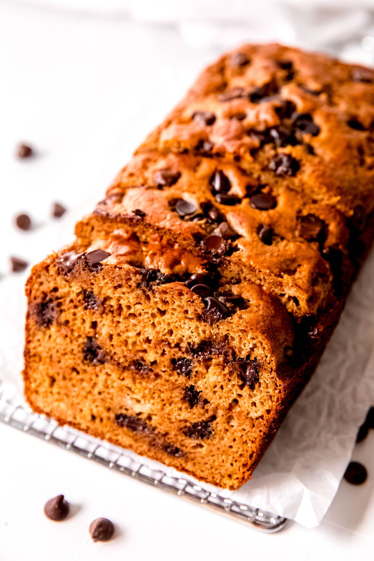 45 degree angle downward shot looking at a loaf of protein powder banana bread with chocolate chips and nuts on a cooling rack with chocolate chips scattered on the white table around it.