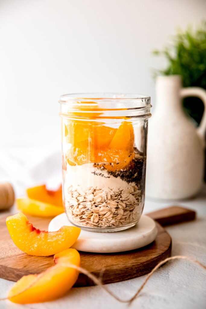 oats, protein powder, chia seeds, sliced peaches, greek yogurt, peach syrup, and vanilla extract added to a mason jar for making no-cook overnight oats.