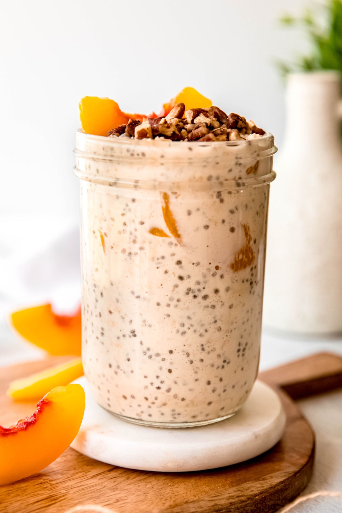 side on shot of a serving of peach overnight oats with protein powder in a mason jar on a white coaster on a wooden cheese board with slices of peaches scattered around.