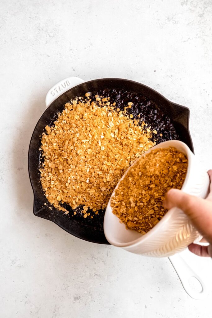 action shot of a hand sprinkling the oatmeal crisp topping evenly across the blackberry filling in the pan.