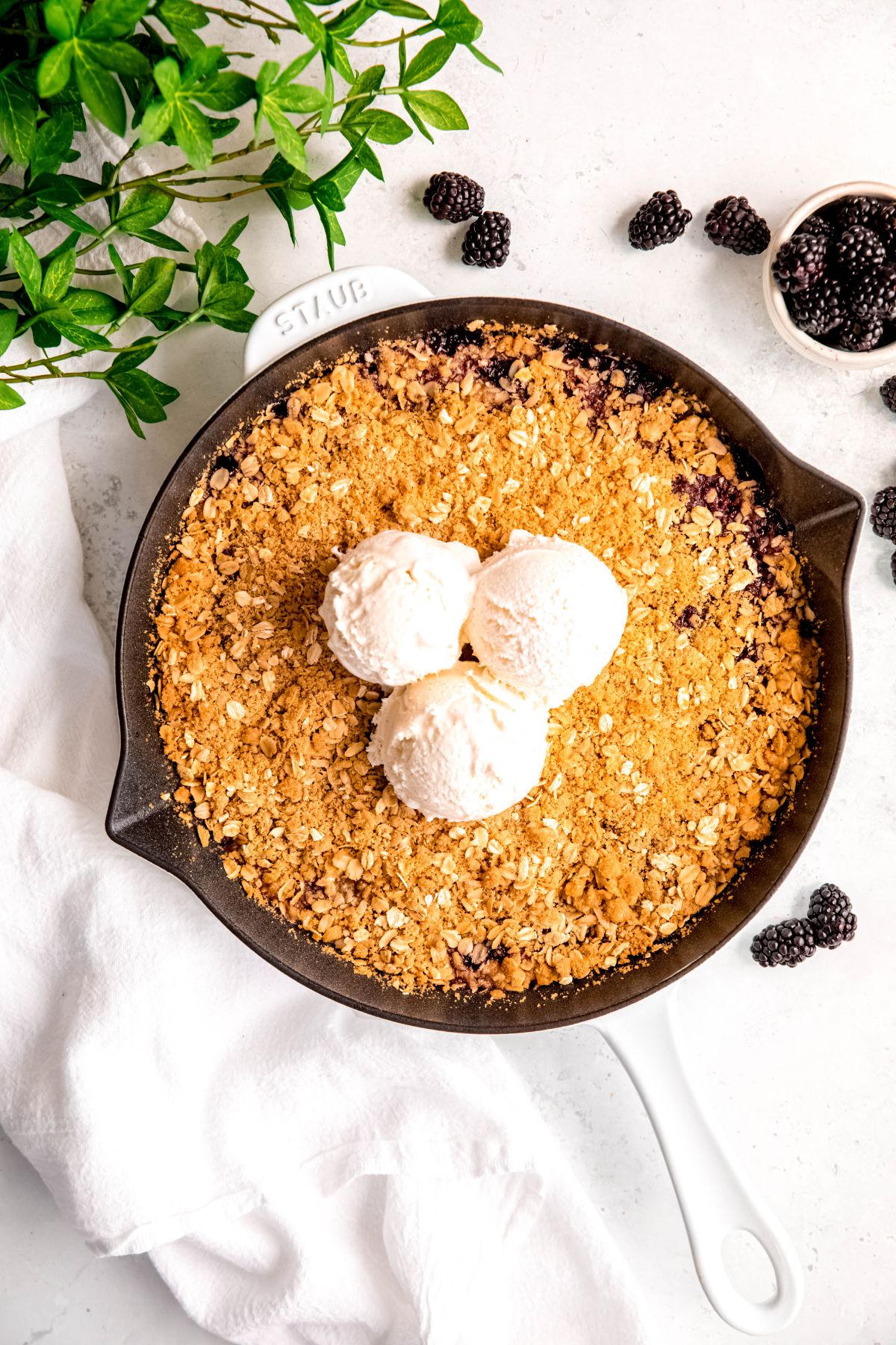 overhead shot of a fresh blackberry crisp in a white cast iron skillet tipped with 3 scoops of vanilla ice cream on a white table with a bowl of fresh blackberries.