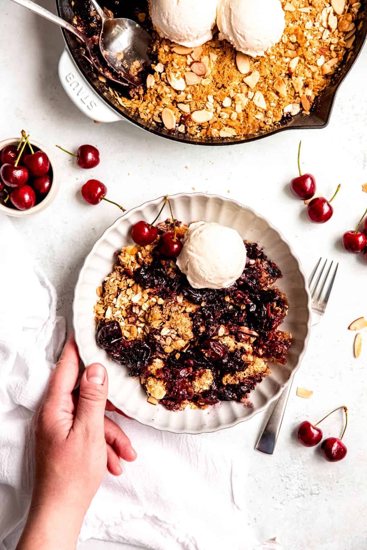 An overhead shot of a hand holding a bowl of cherry cobbler with a scoop of ice cream and fresh cherries.