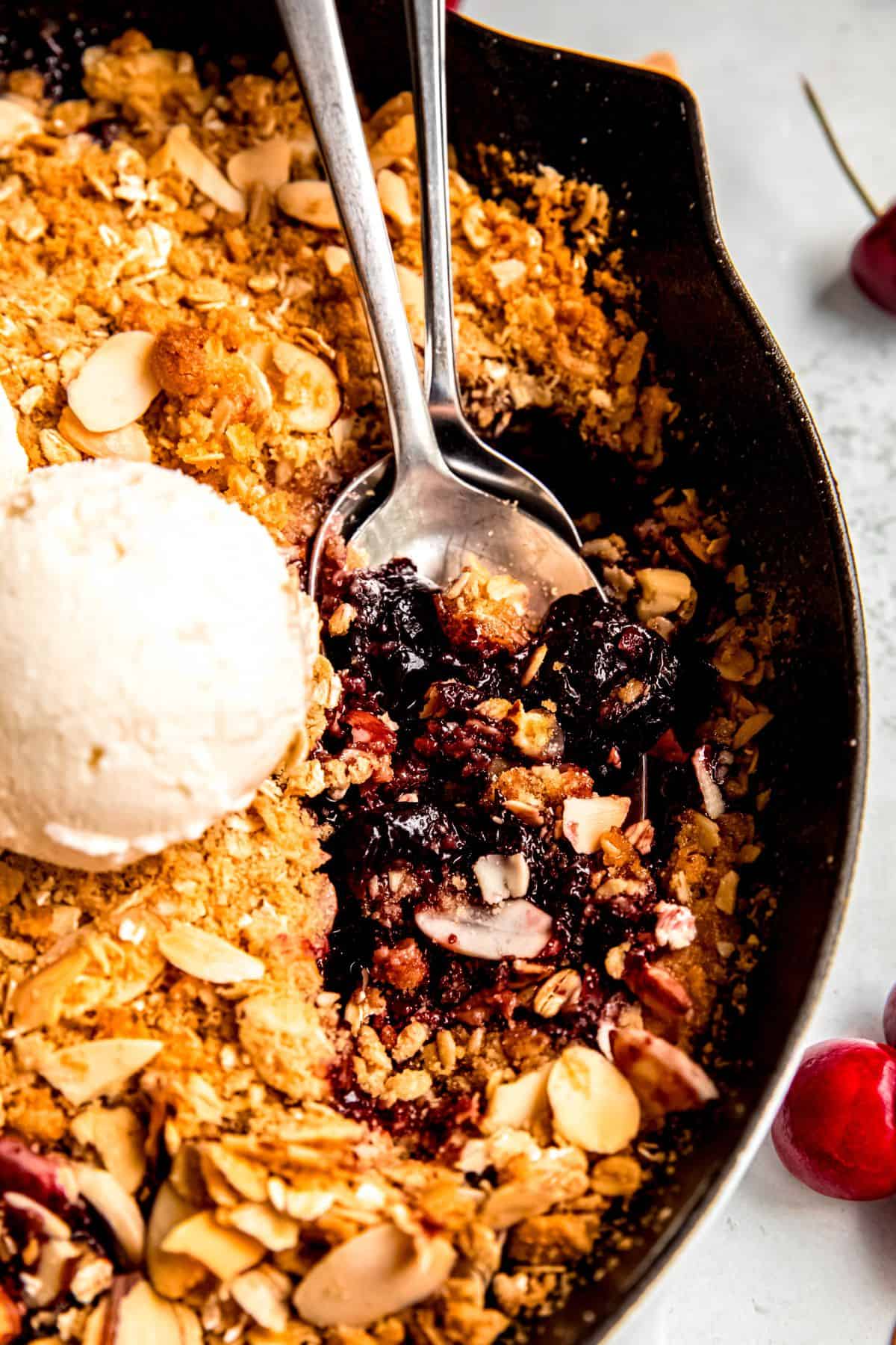 A close-up shot of cherry crisp with two spoons in it and a scoop of ice cream.