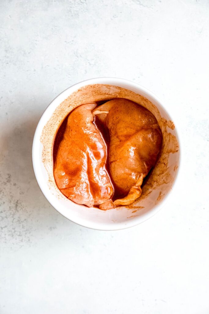 raw chicken breasts in a white mixing bowl with the hot honey marinade poured on top and squished around.