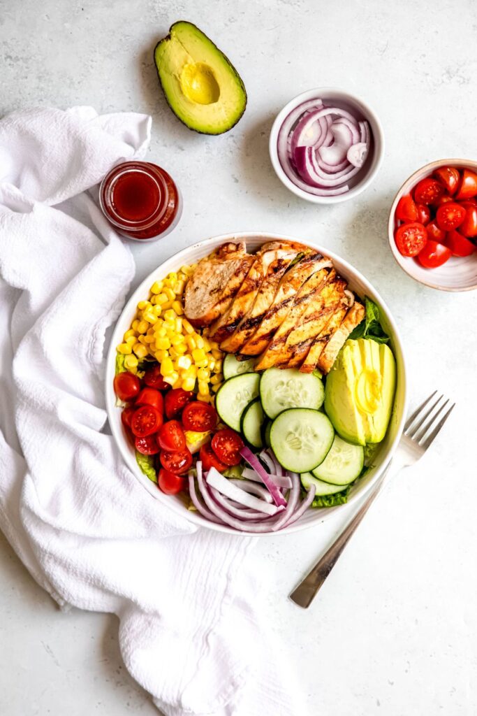 sliced grilled hot honey chicken added to the salad; there's extra hot honey vinaigrette, a halved avocado, and two ramekins filled with red onion slices and cherry tomato halves across the top of the frame, a white linen to the left of the bowl, and a silver fork to the right.