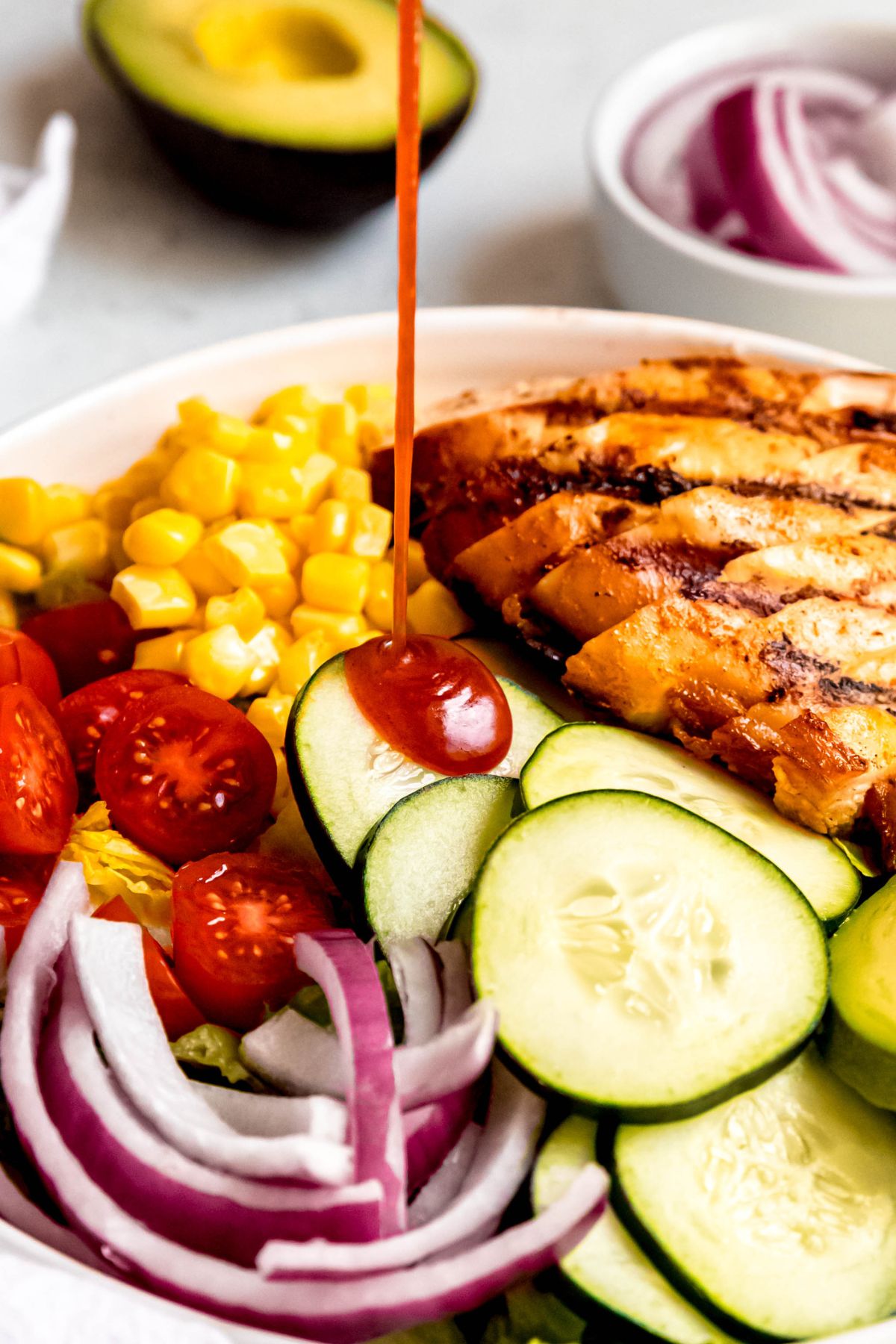 action shot of hot honey salad dressing being drizzled atop a plate with sliced grilled chicken, cucumbers, red onions, cherry tomatoes, and corn.