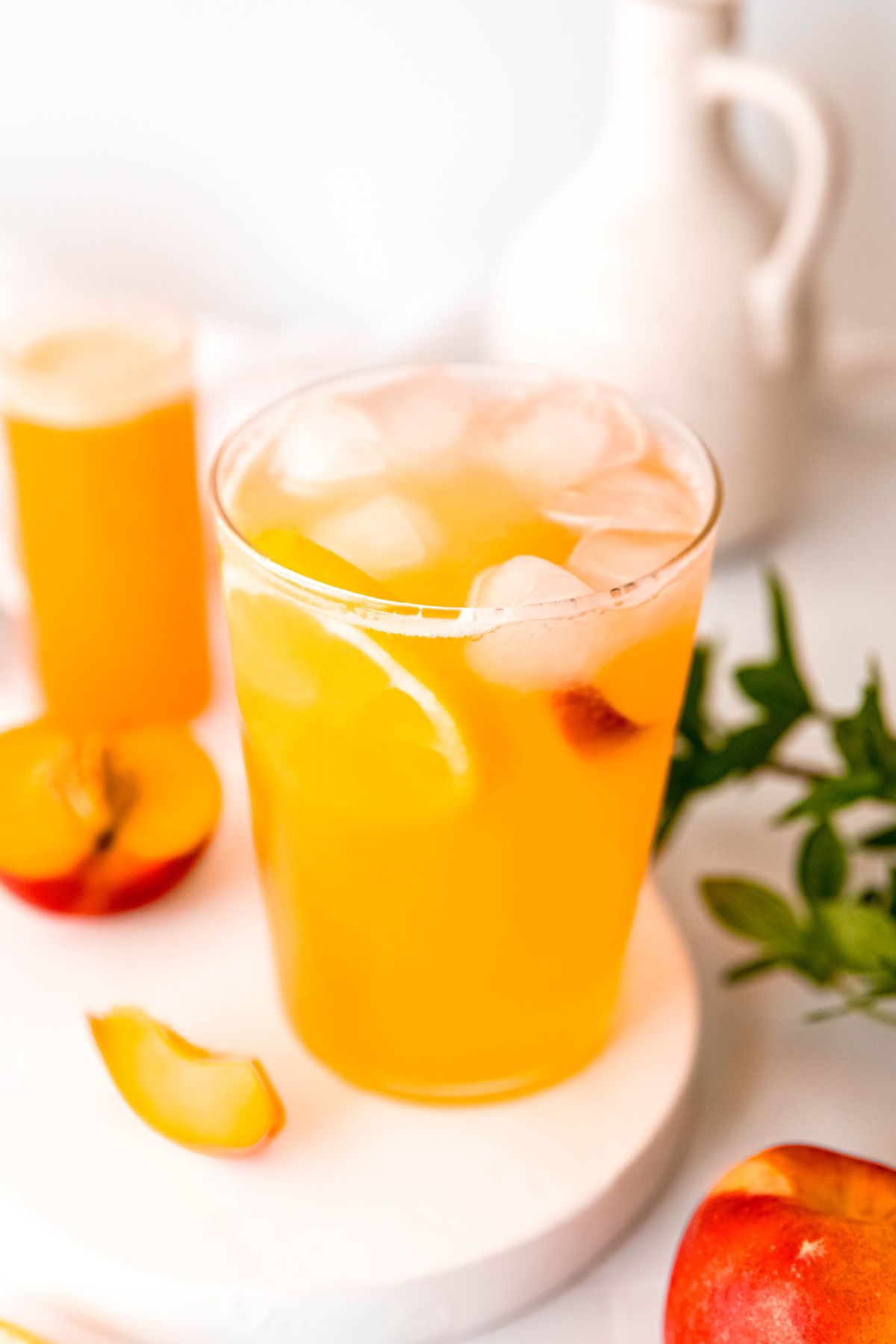 45 degree downward angle shot into a glass of 3-ingredient nectarine lemonade on a white pedestal on a white table with a green houseplant, a collins glass of nectarine drink syrup, and fresh nectarines on the table.