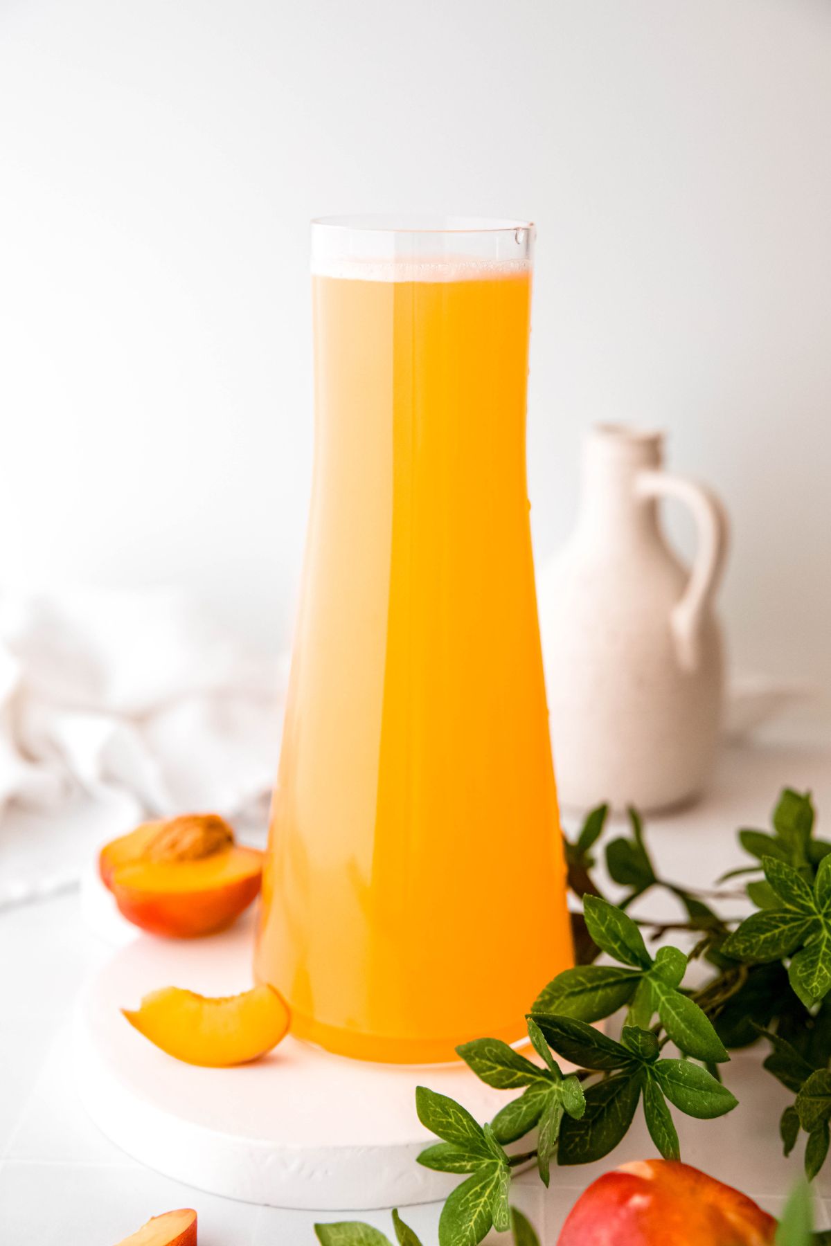 hourglass shaped handle-less glass pitcher filled with homemade nectarine lemonade on a white table with a white ceramic jug and a halves nectarine blurred out in the back of the shot, and a few branches of a green houseplant and a whole nectarine in the front right corner.