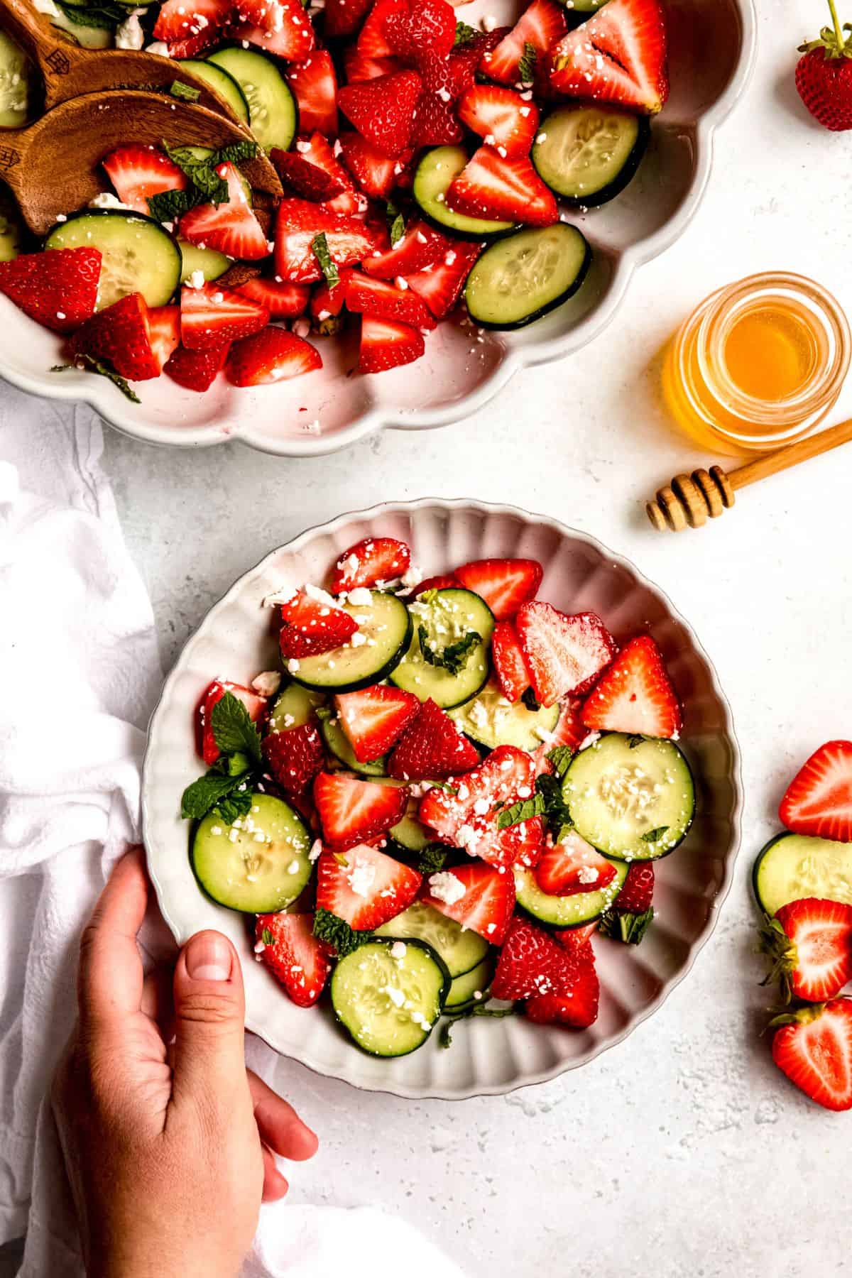 hand grabbing a white scalloped salad bowl with a serving of strawberry cucumber salad with the remaining cucumber strawberry salad in the larger serving bowl in the top of the frame.