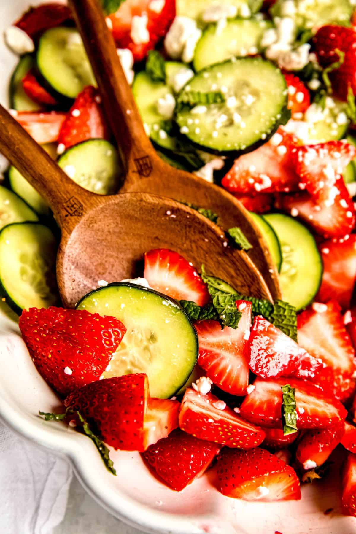 closeup shot of dark wooden spoons in a bowl of cucumber strawberry salad after tossing.
