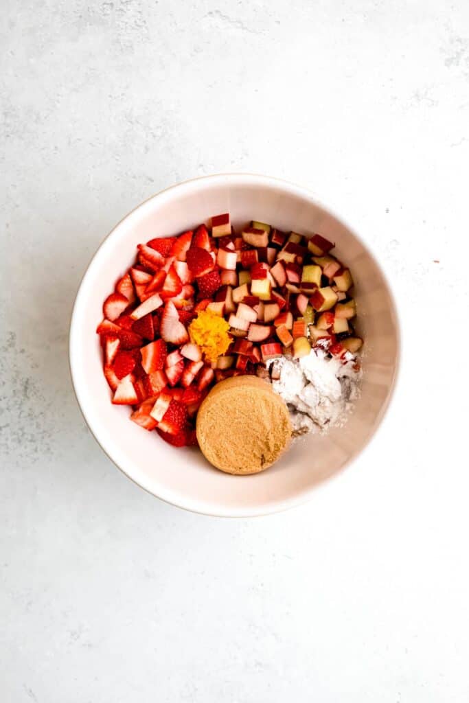 An overhead shot of a mixing bowl with chopped strawberries, rhubarb, orange zest, brown sugar, and cornstarch.