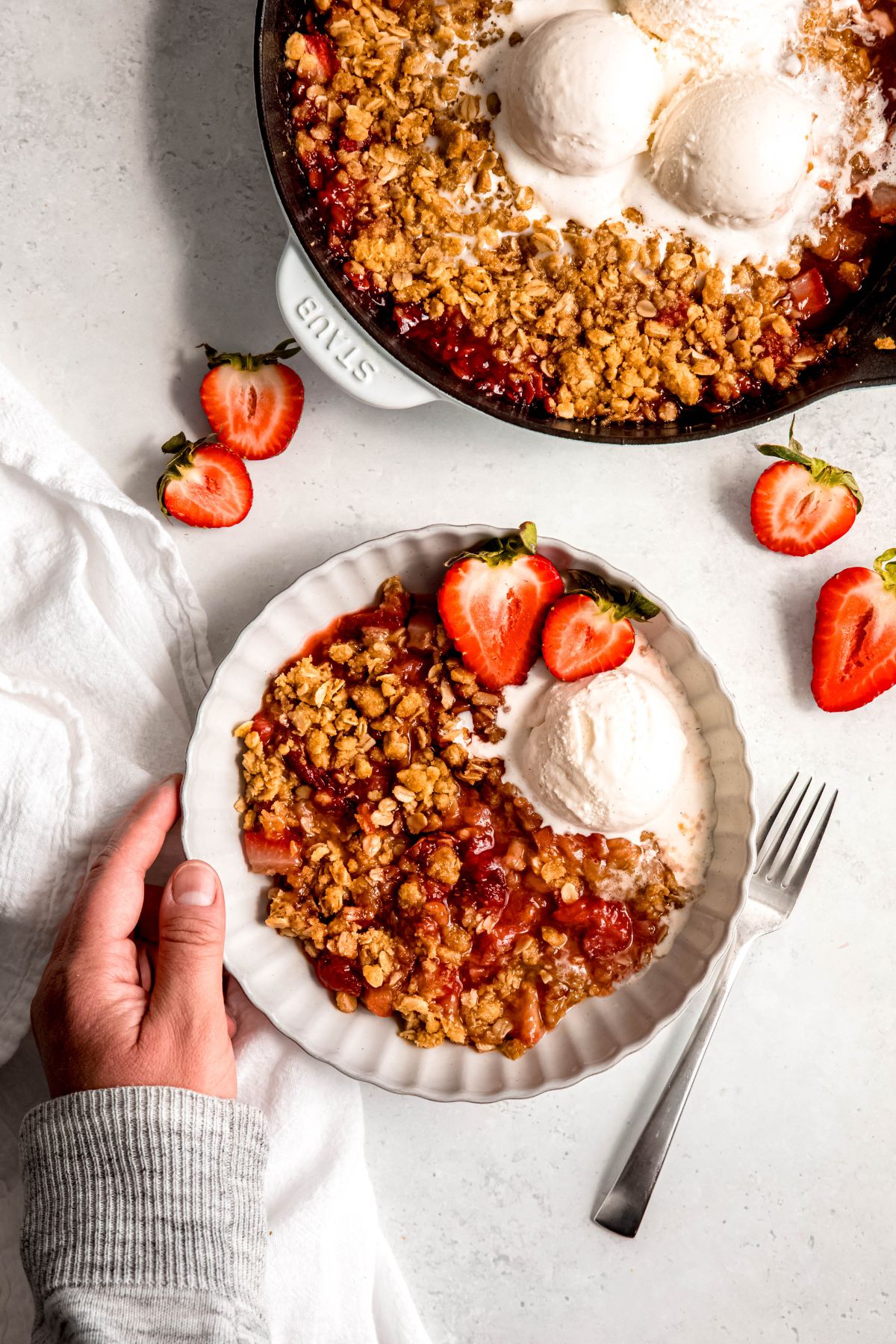 An overhead shot of a hand holding a bowl of strawberry rhubarb crisp next to a skillet of the crisp.
