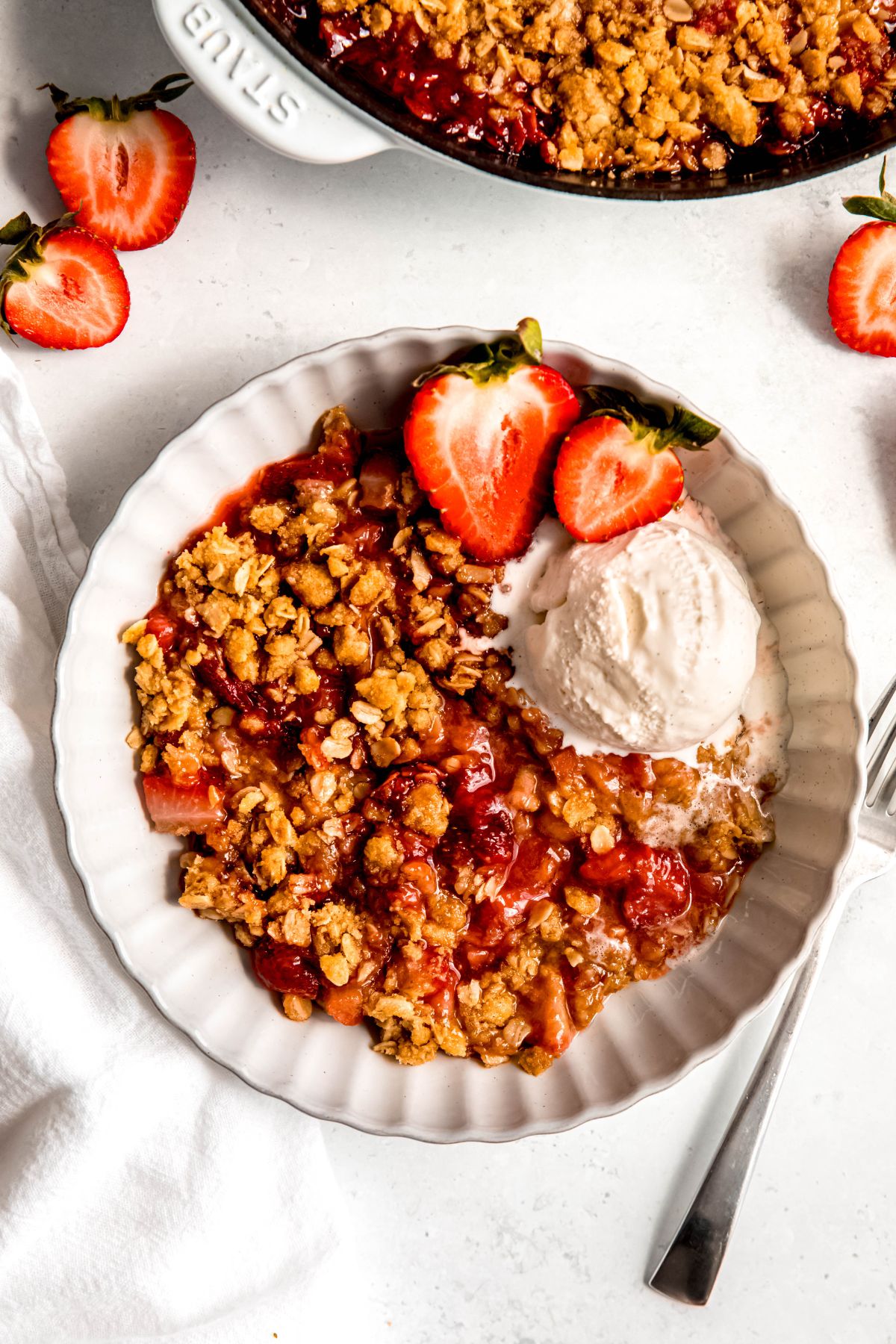 An overhead shot of a large serving of strawberry rhubarb crisp with a scoop of ice cream and a halved berry.