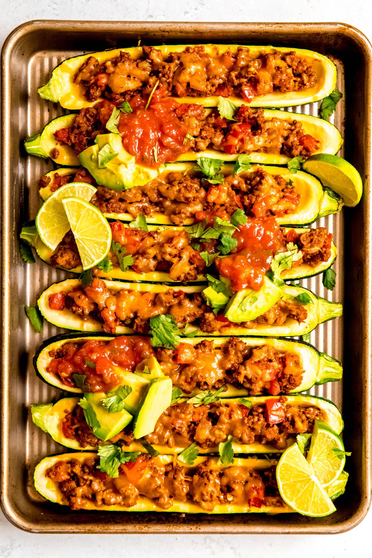 An overhead shot of a pan of taco zucchini boats topped with avocado, limes, and cilantro.