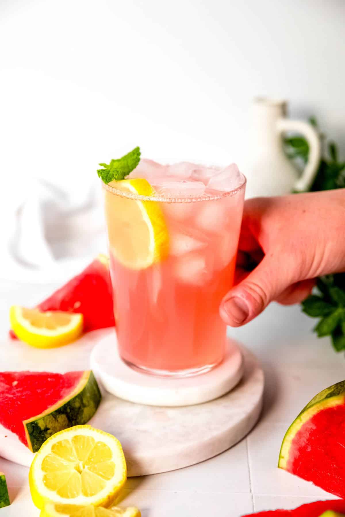 hand grabbing a pint glass of watermelon lemonade from a white coaster on a white table with brightly colored wedges of fresh watermelon and fresh lemon slices scattered around.