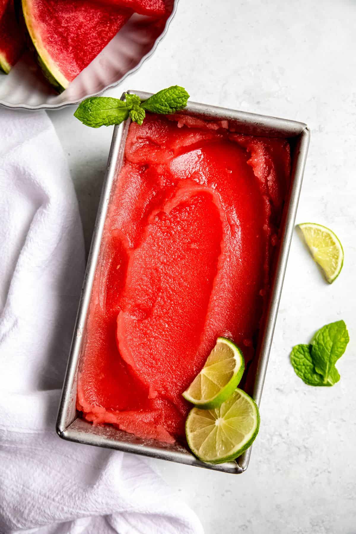 homemade watermelon sorbet in a metal loaf din on a white table with the top showing that a scoop has been taken out; it's garnished with a mint sprig in the top left corner and lime wheels in the bottom right corner.