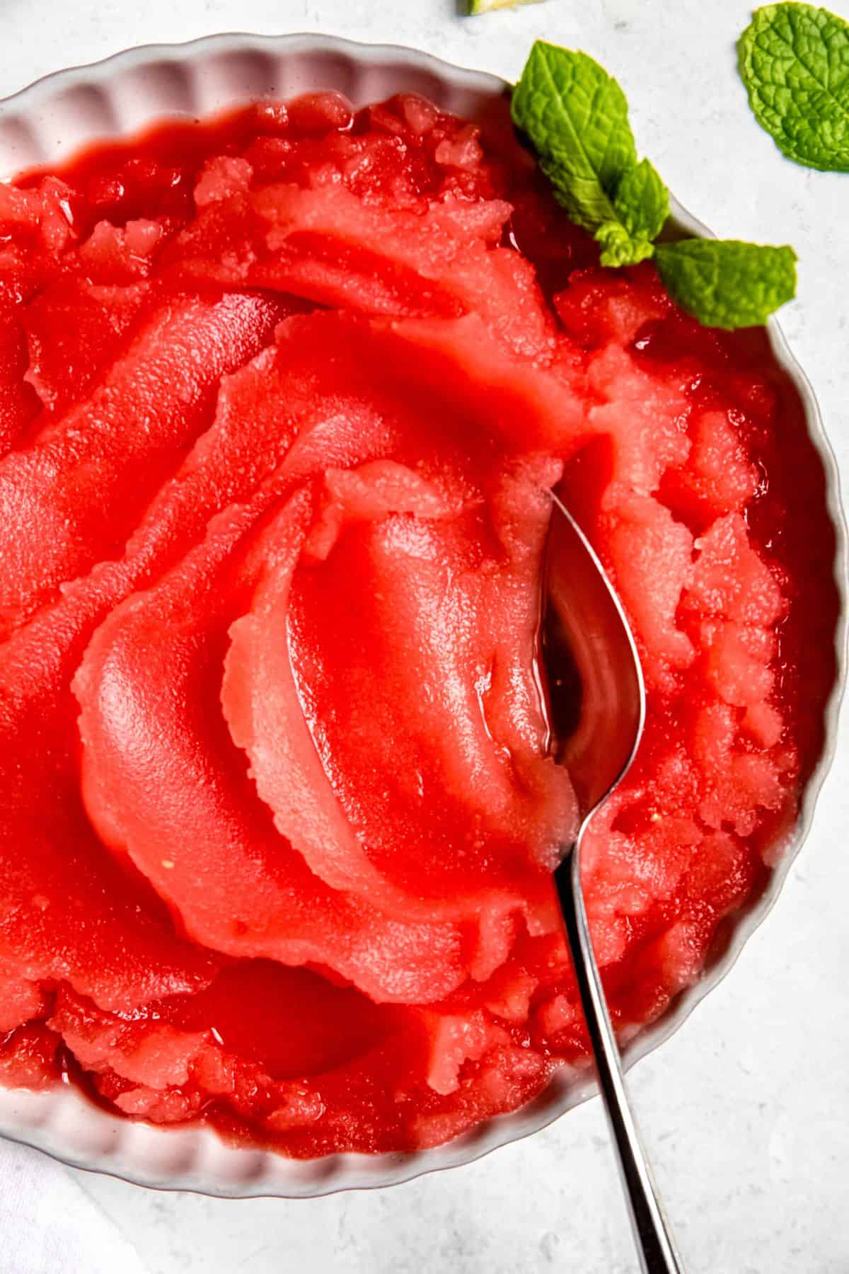closeup overhead shot of a silver spoon taking a bite of fresh watermelon sorbet from a white bowl.