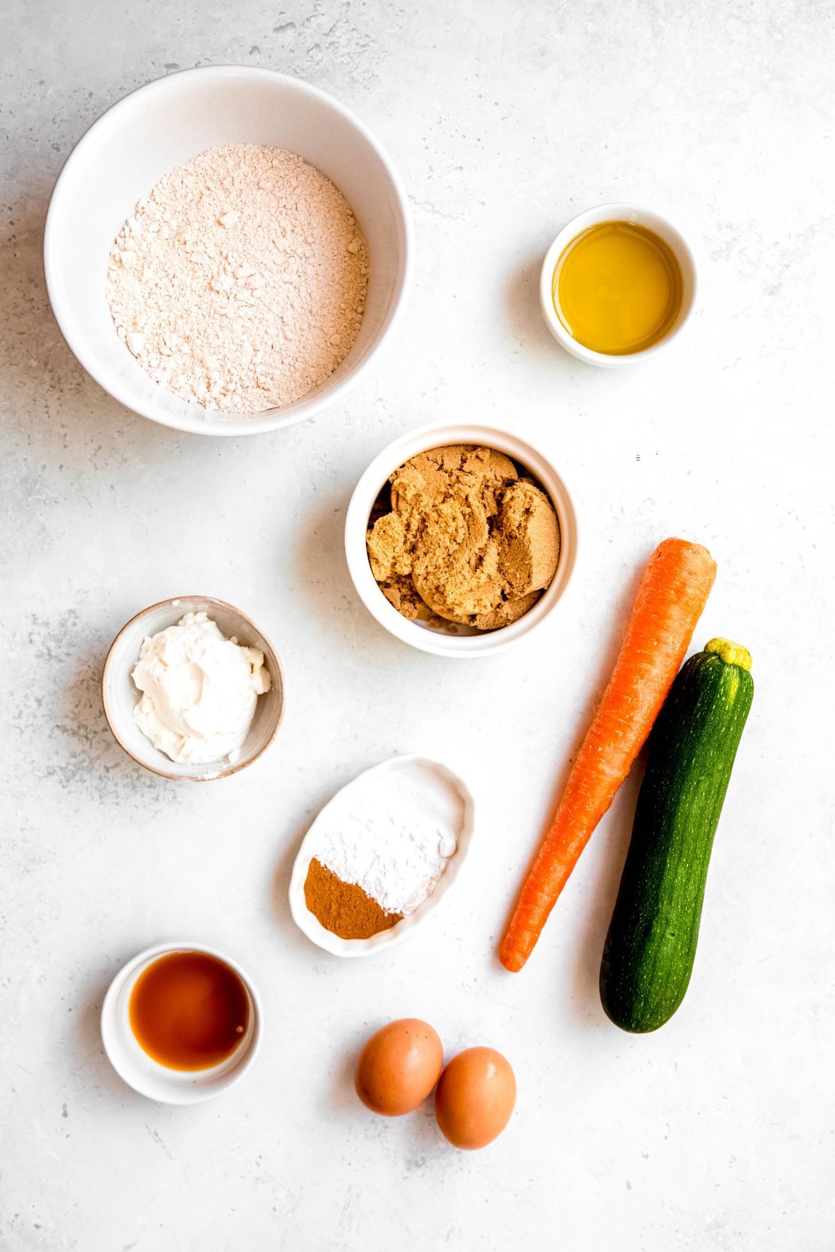 ingredients for making healthy carrot and zucchini muffins measured out on a white table.