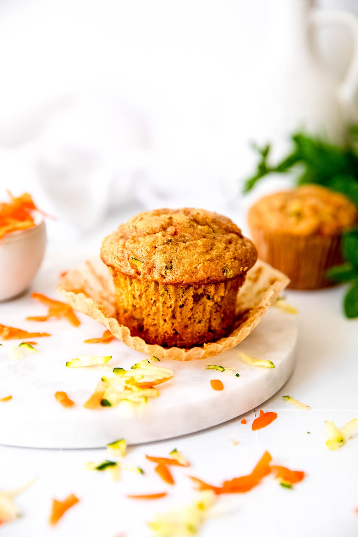 table with a couple of healthy zucchini carrot muffins; one has been partially unwrapped and there are shreds of fresh zucchini and carrot scattered around.