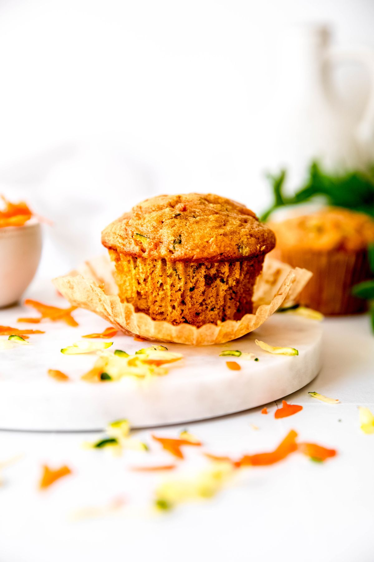 carrot zucchini muffin in a kraft paper wrapper that's been partially unwrapped on a white table with shreds of fresh zucchini and carrots scattered around.