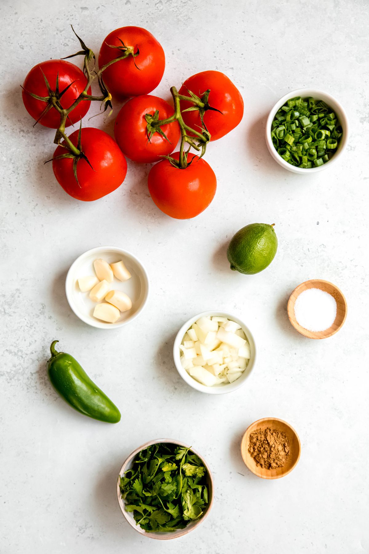 fresh tomatoes, diced scallions, diced onion, fresh lime, fresh jalapeño, fresh garlic cloves, salt, cumin, and fresh cilantro measured out for making homemade fresh tomato salsa recipe.