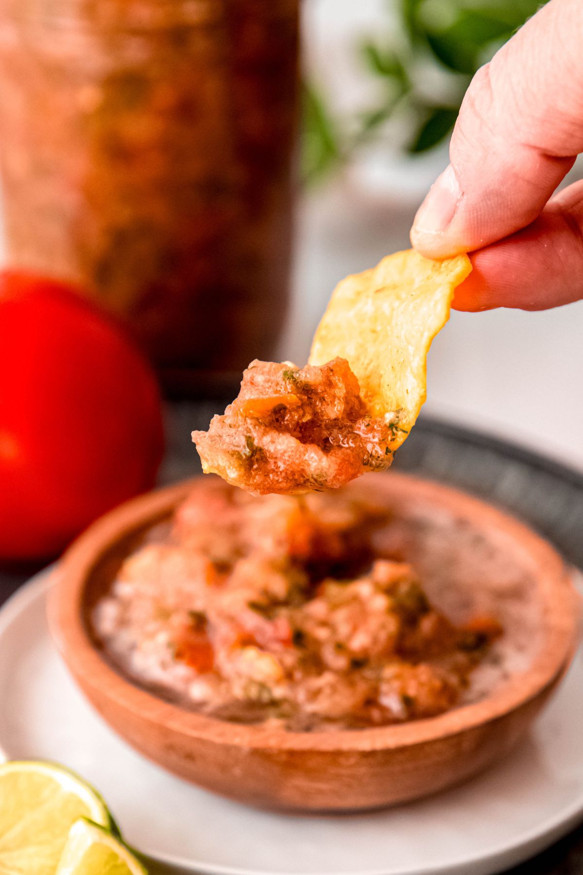 hand with a tortilla chip that has been dipped in a bowl of chunky fresh restaurant style salsa with the rest of the bowl and fresh tomatoes blurred out in the background.