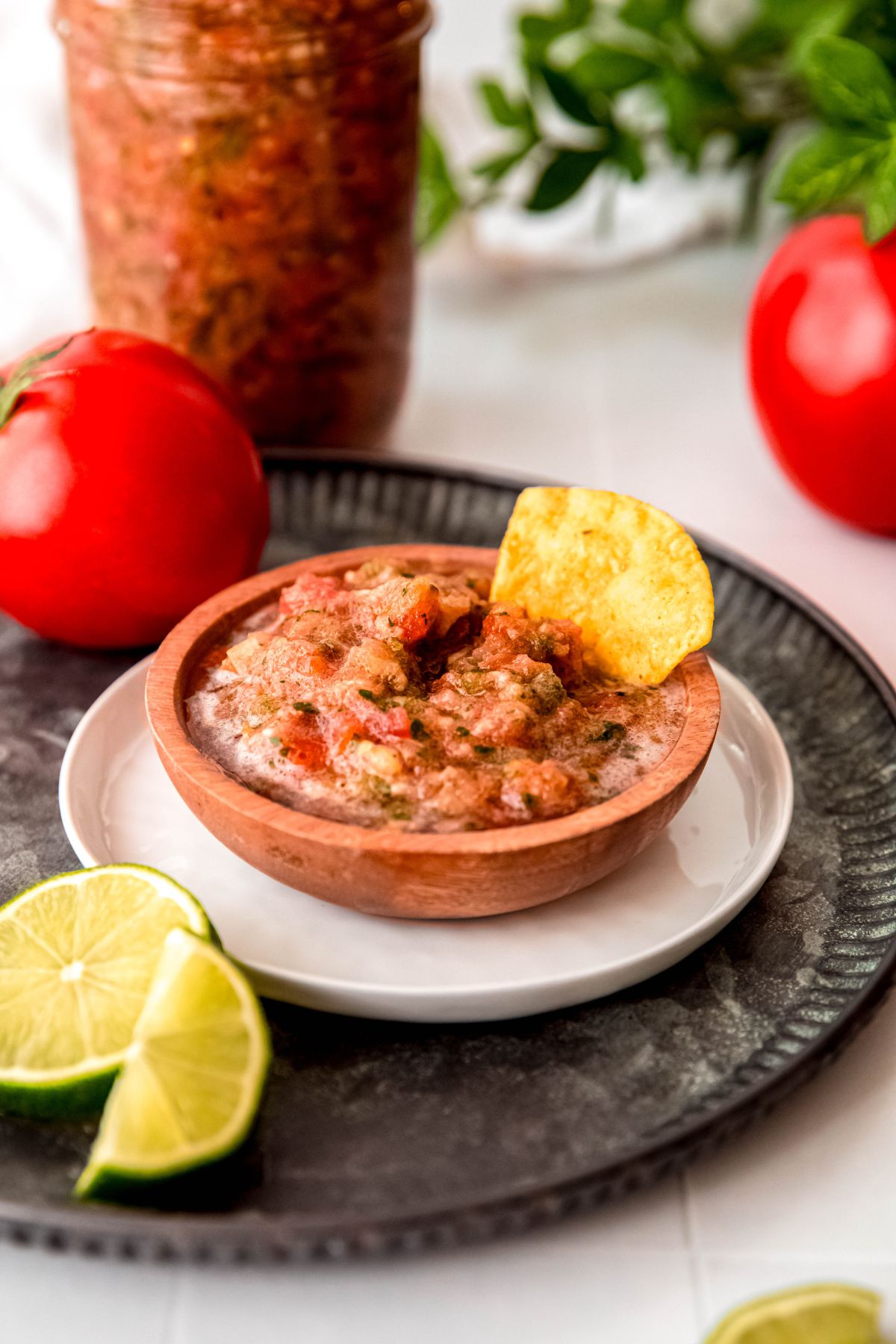 wooden bowl of fresh tomato salsa on a white plate on a hammered black metal serving tray with a single chip dipped in; on the tray there's a halved lime and whole fresh tomato, and on the white table in the background there's a mason jar with the rest of the batch of salsa and another fresh tomato.