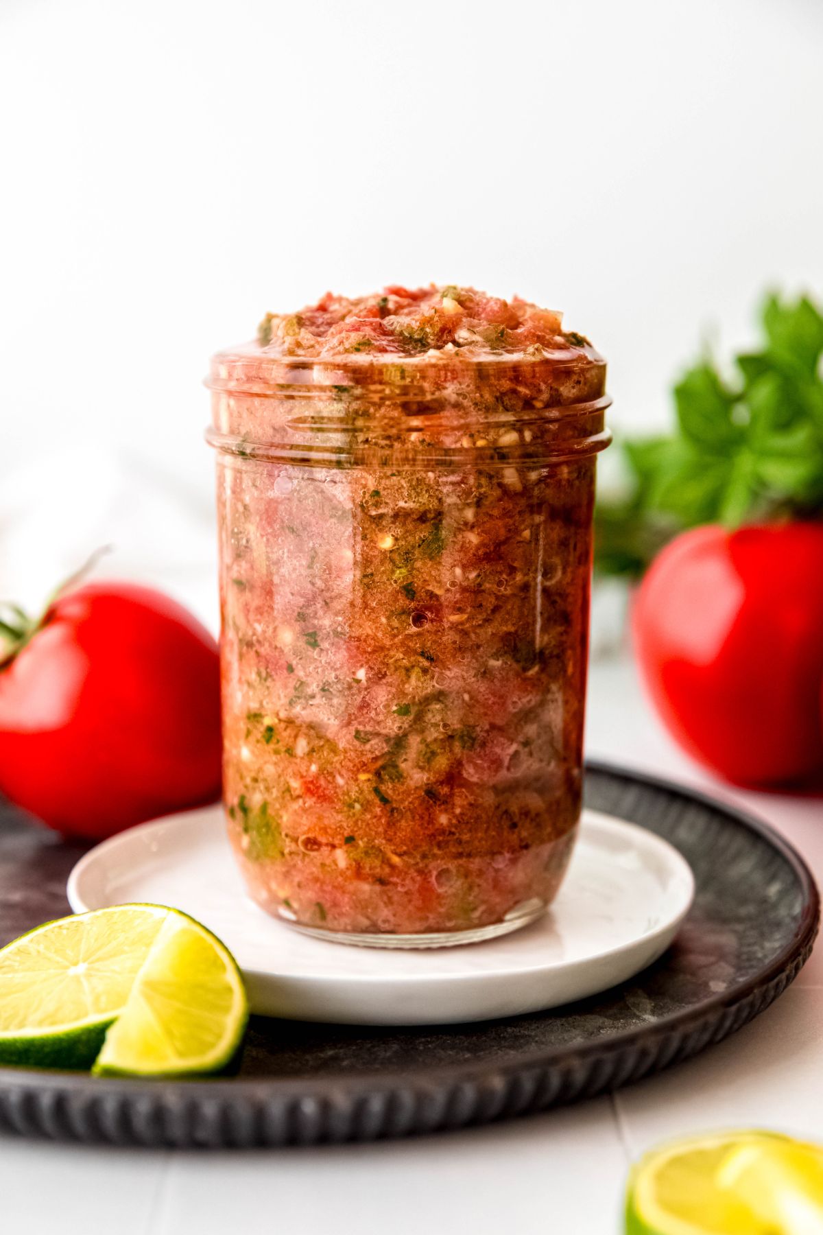 mason jar brimming with restaurant style tomato salsa on a white plate on a black metal serving tray on a white table with fresh cilantro and tomatoes blurred out in the background and a fresh lime in the foreground.
