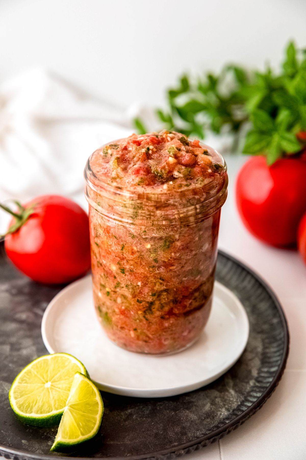 jar of homemade restaurant-style salsa on a white plate on a black serving platter on a white table with fresh tomatoes, fresh lime, and fresh cilantro scattered around.