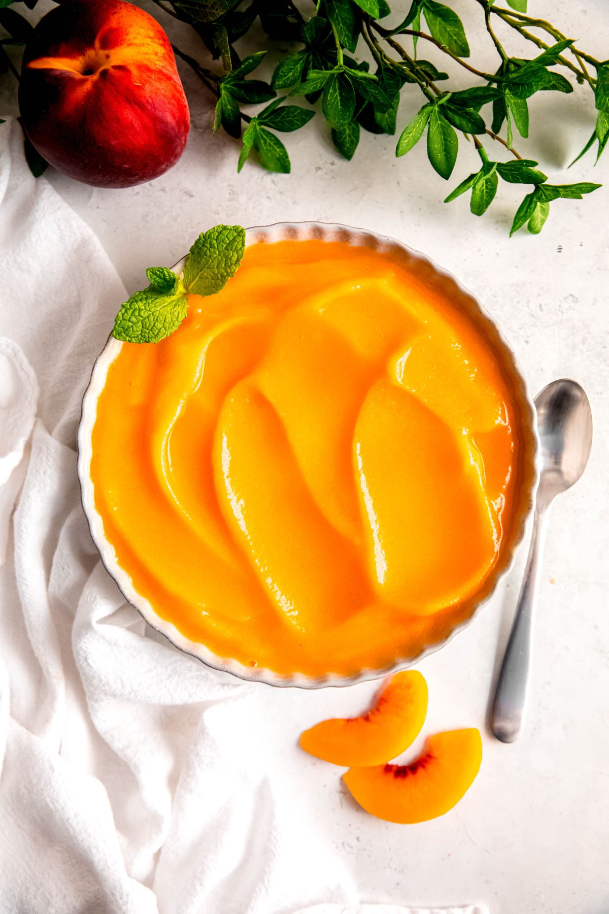 overhead shot of a bowl of homemade peach sorbet garnished with a sprig of mint on a white table with a white linen, silver spoon, two slices of fresh peach, a whole peach, and sprigs from a green plant surrounding the bowl.