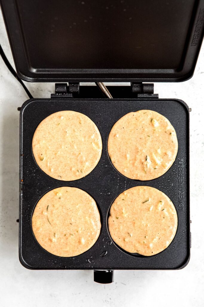 electric pancake skillet with 4 wells filled with zucchini pancake batter.