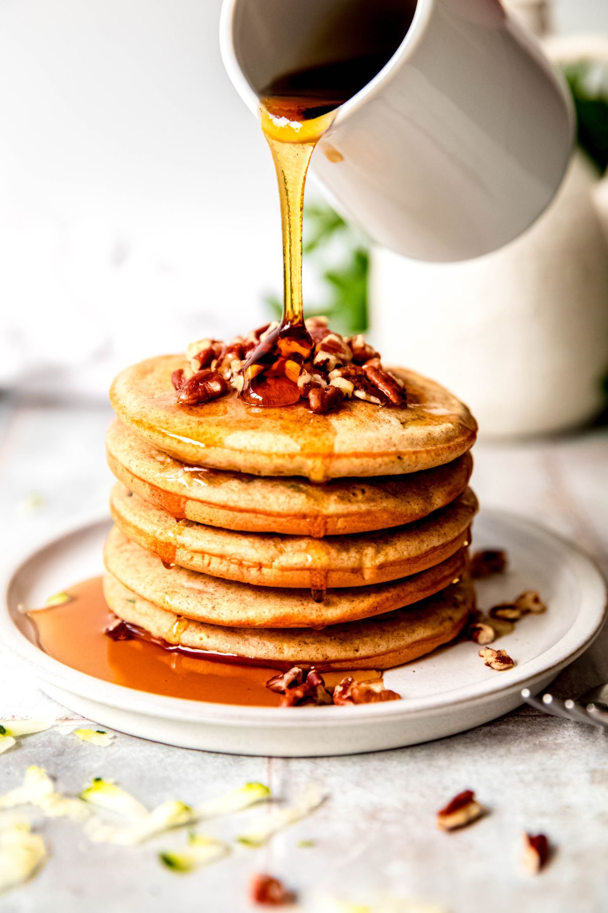 action shot of maple syrup being drizzled on a short stack of high protein zucchini bread pancakes topped with pecans.